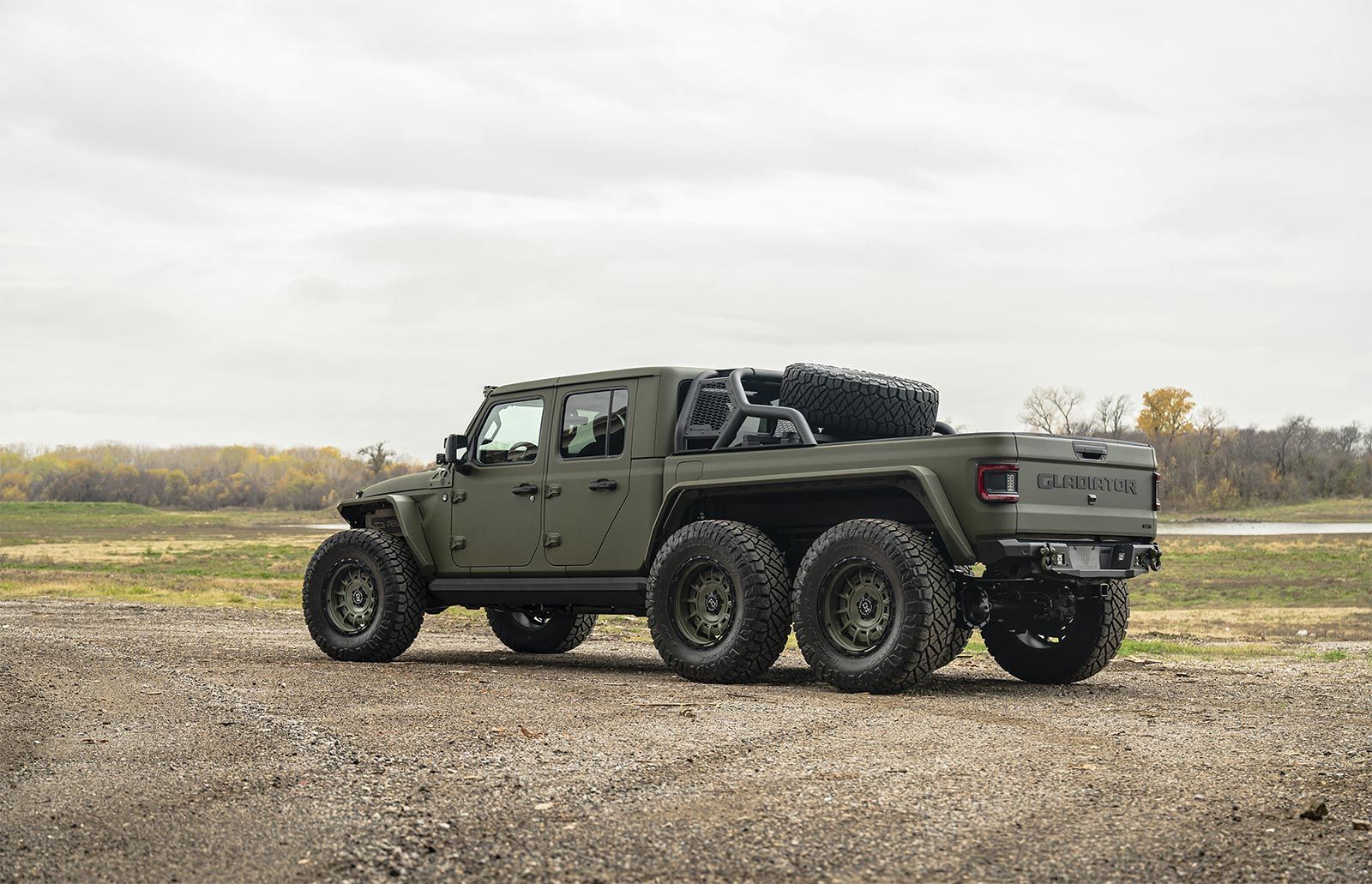 A green jeep with six wheels is parked in a dirt field.