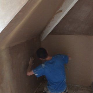 Person plastering a wall in a room with a sloped ceiling and wooden support beam, using plaster and a trowel.