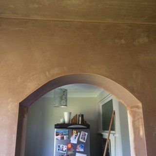 Brown plastered wall with arched doorway, revealing a kitchen with refrigerator.