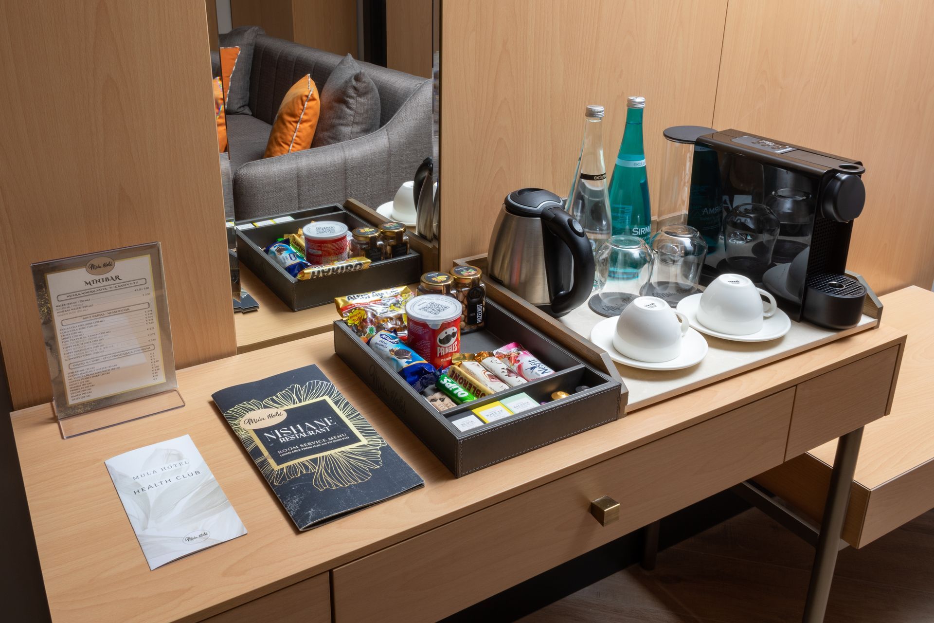 Mula Hotel | Hotel room coffee station with snacks, water bottles, cups, and a coffee maker on a desk.