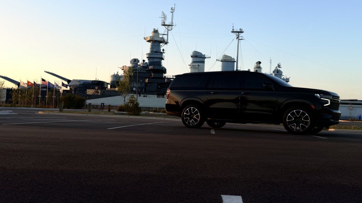 A black SUV parked on an asphalt lot in front of a battleship at sunset.