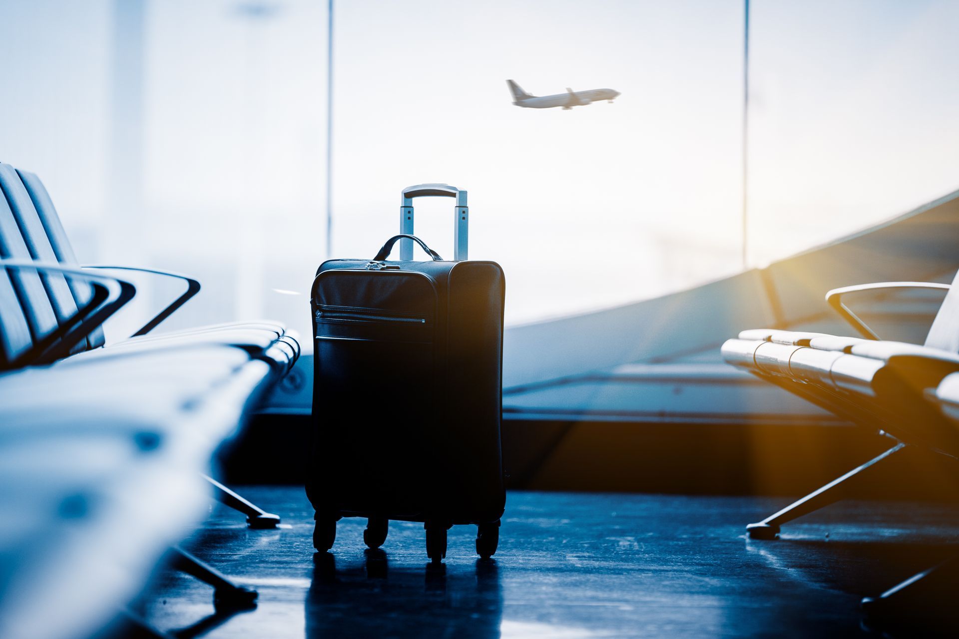 A black suitcase standing in an empty airport terminal waiting area with an airplane visible through the window.