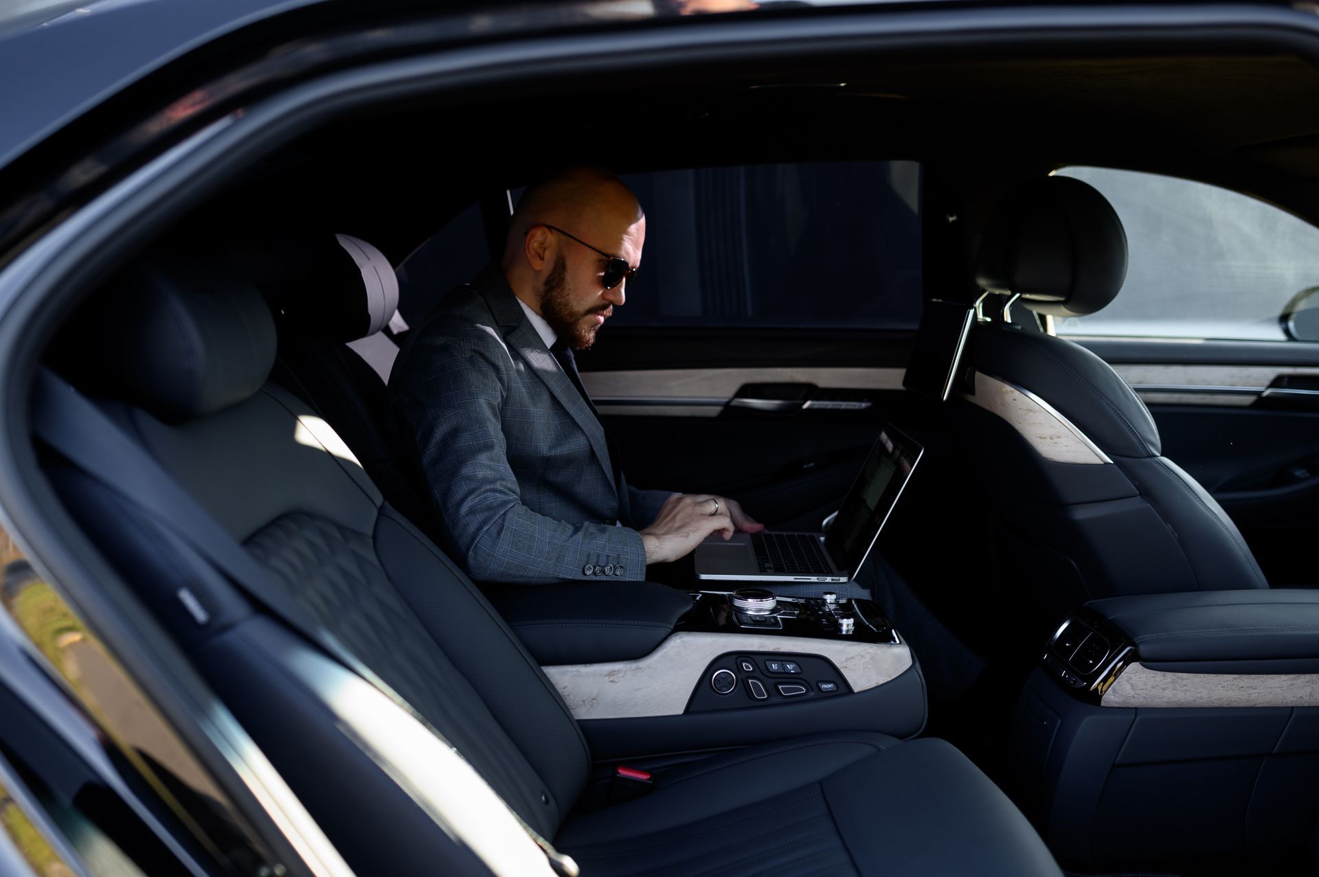 A person in a suit and sunglasses works on a laptop in the back seat of a luxury car with black leather interior.