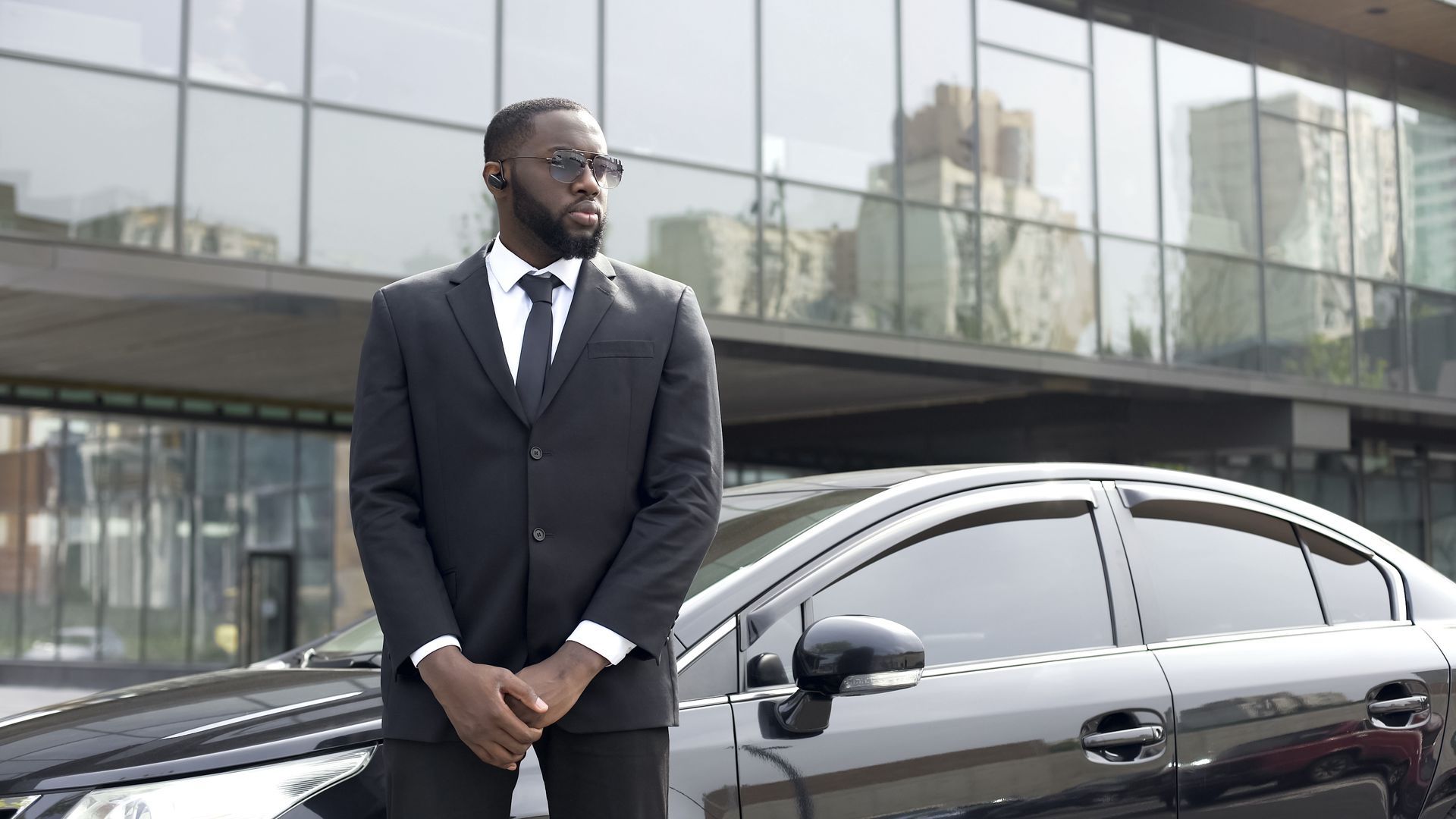 A professional in a suit and tie standing in front of a modern glass building and a dark car, hands clasped.