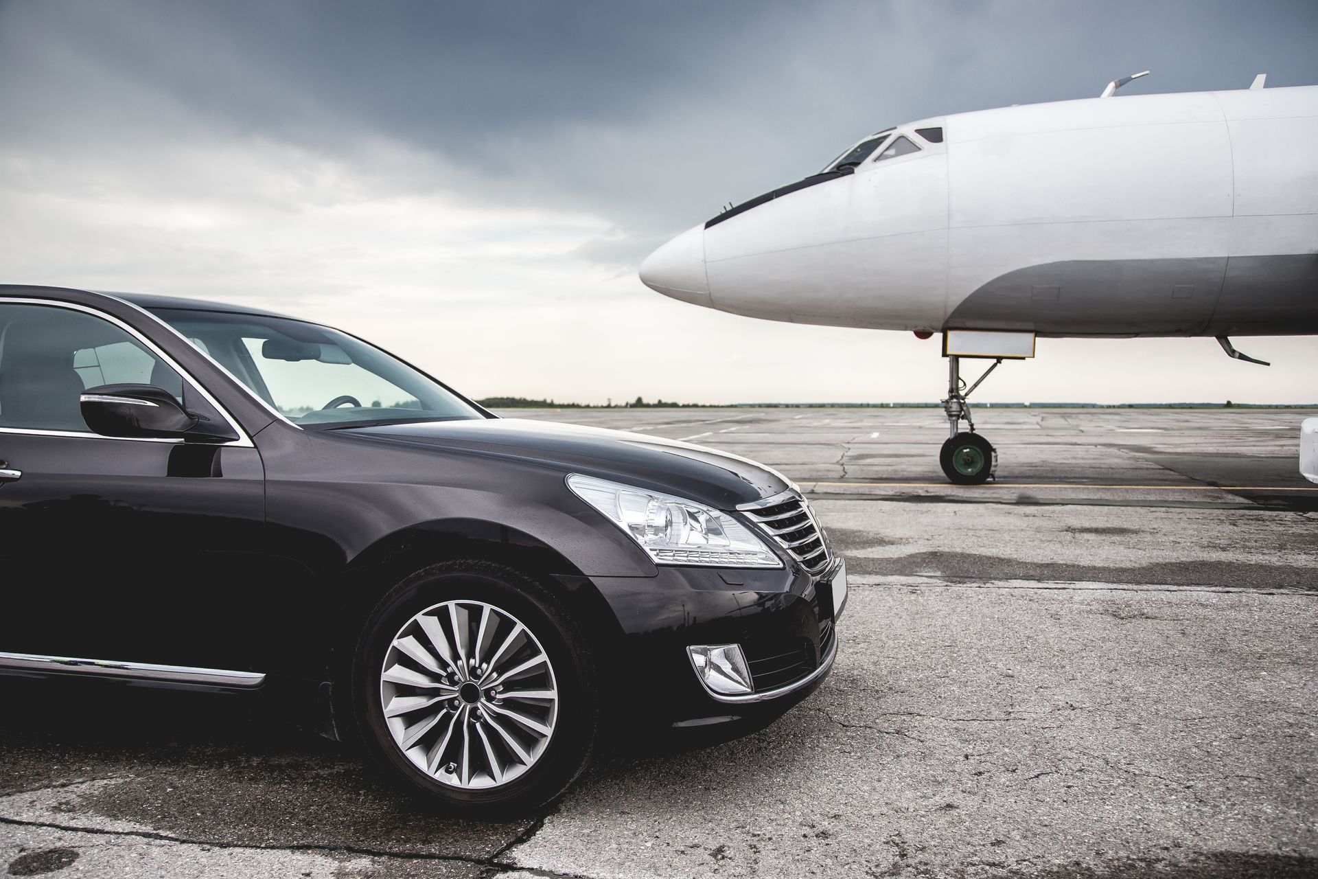 A dark sedan parked on an airport tarmac next to the nose of a large white airplane under a cloudy sky.