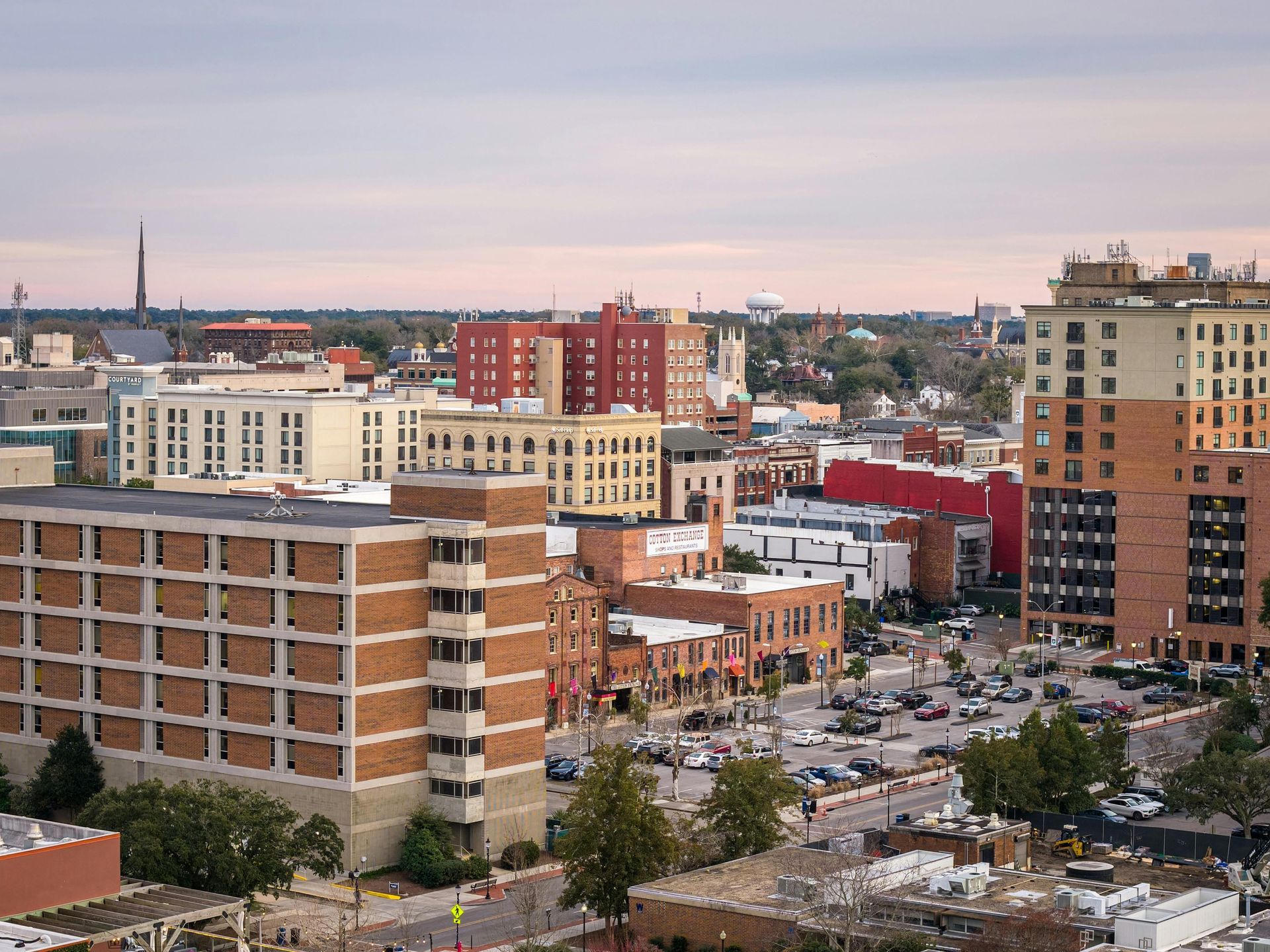 Aerial view of a city downtown with brick and beige buildings, parking lots, and trees under a dusk sky.