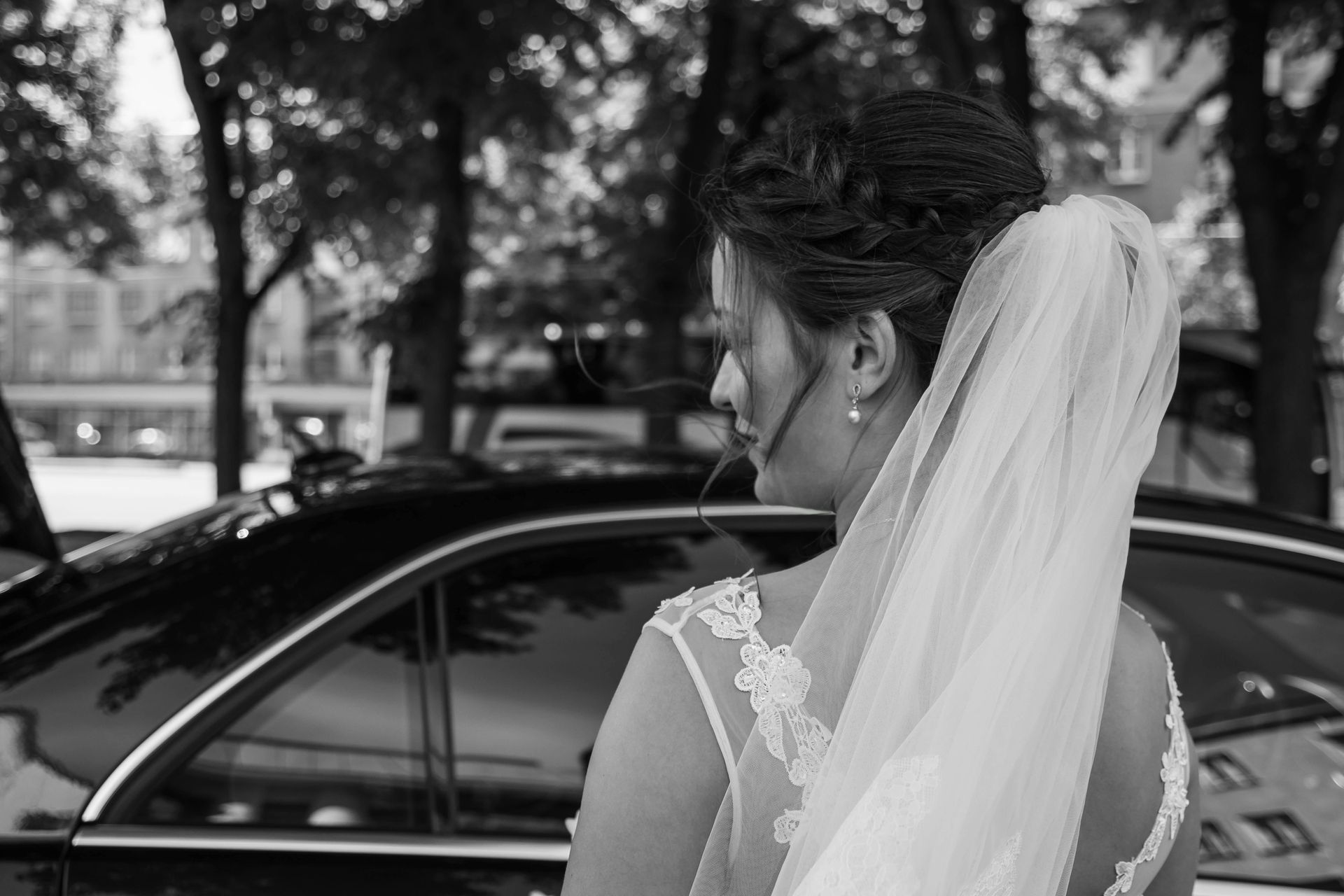 A bride in a lace wedding dress and veil stands looking toward a black car outdoors, captured in black and white.