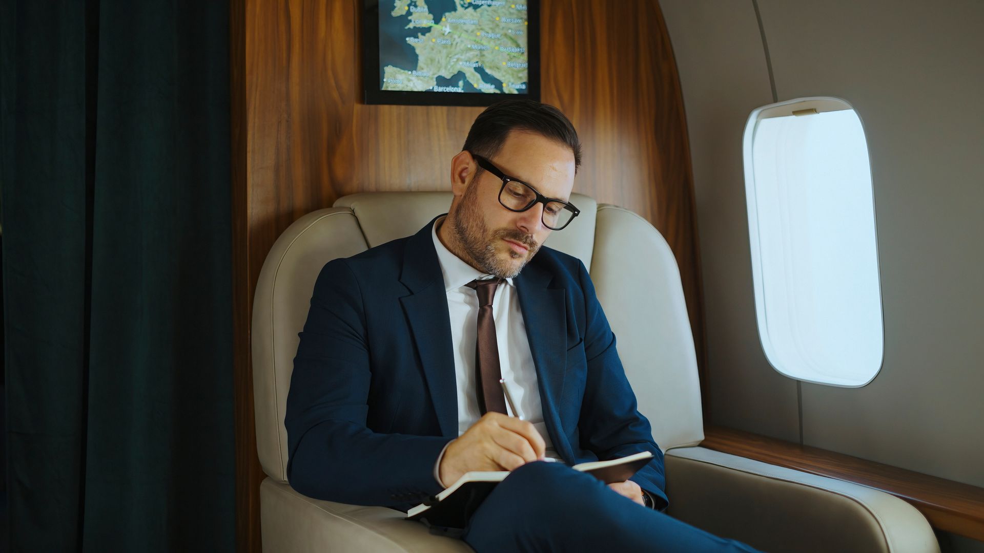 A person in a suit sits in a private airplane seat, writing in a notebook near a window.
