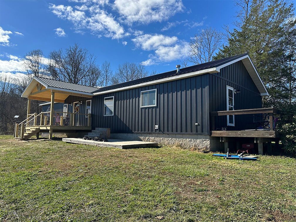 A black mobile home with a porch and stairs is sitting in the middle of a grassy field.