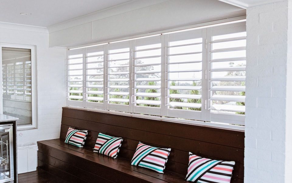 A Wooden Bench with Colorful Pillows in Front of A Window with White Shutters — New Blinds and Shutters in Maryland, NSW