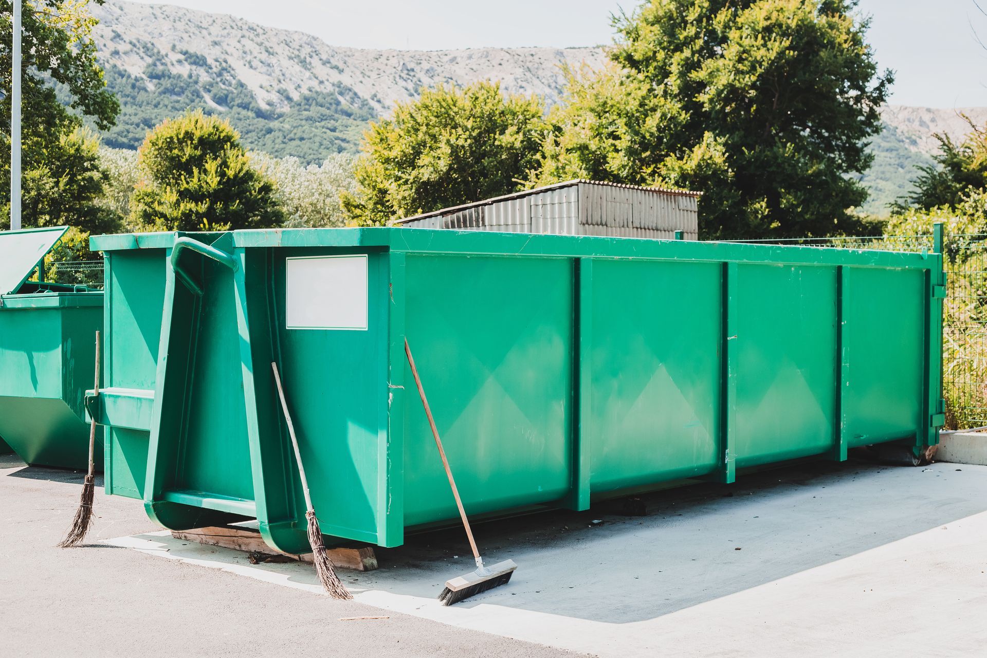 Green industrial dumpster in outdoor setting. Broom leaning against it. Trees and mountains in background.
