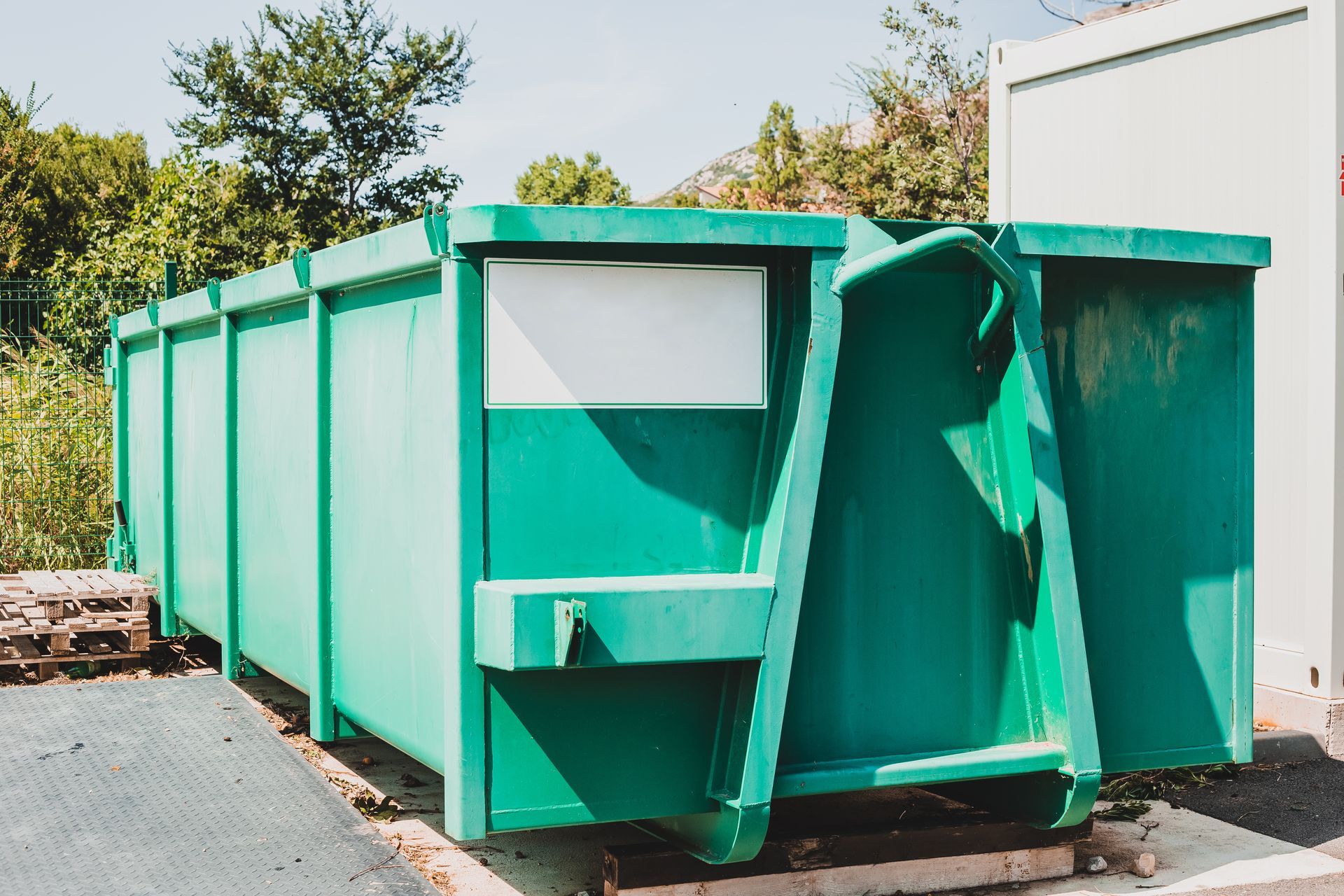 Green dumpster, outdoors, next to a white structure, near trees.