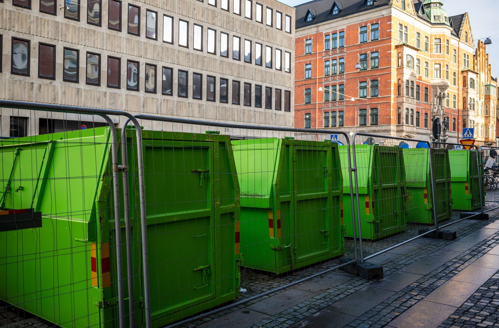Row of green construction containers along a street with buildings.