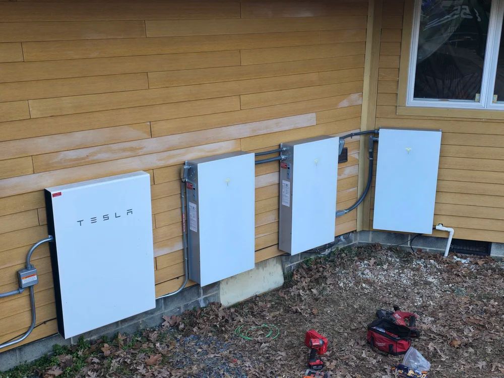 Four white Tesla Powerwalls installed on the exterior of a yellow building, with tools on the ground.