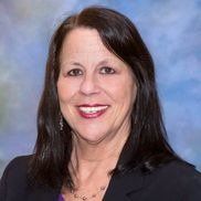 Woman with dark hair smiles, wearing a black blazer and necklace, against a blue and white blurred background.
