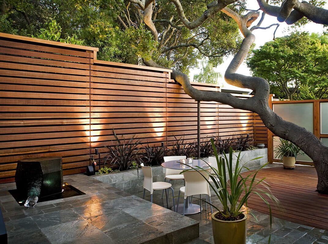 Modern backyard patio with white chairs, glass table, potted plants, and wooden privacy fence at dusk