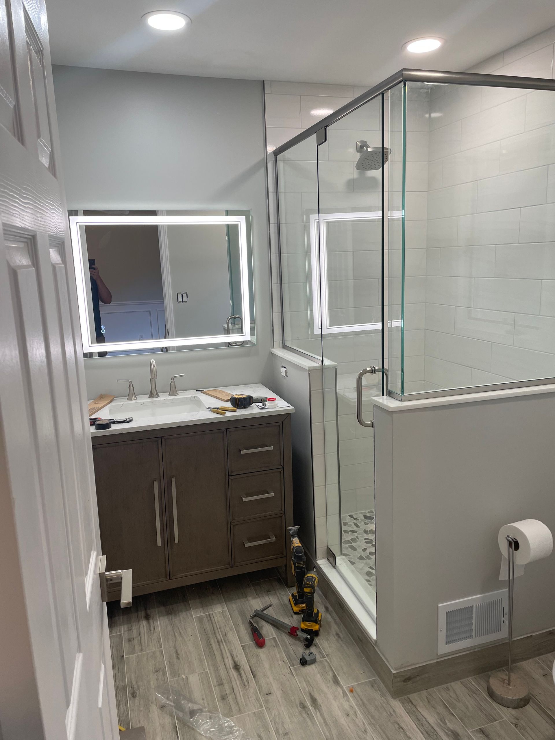 Small bathroom under renovation with vanity, mirror, shower glass, and tools on the floor.