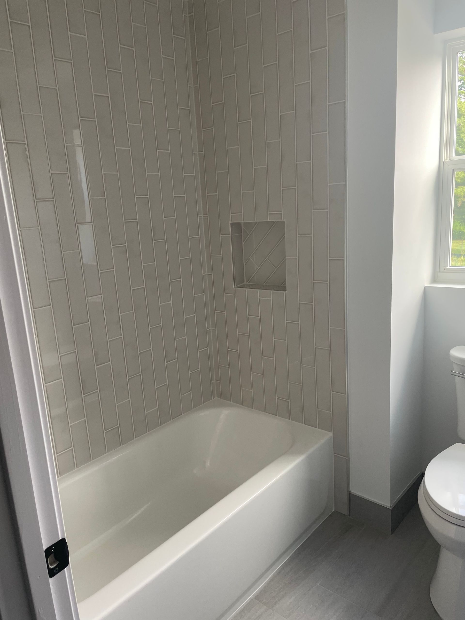 Small bathroom with a white bathtub, tiled shower wall, and part of a toilet beside a window.