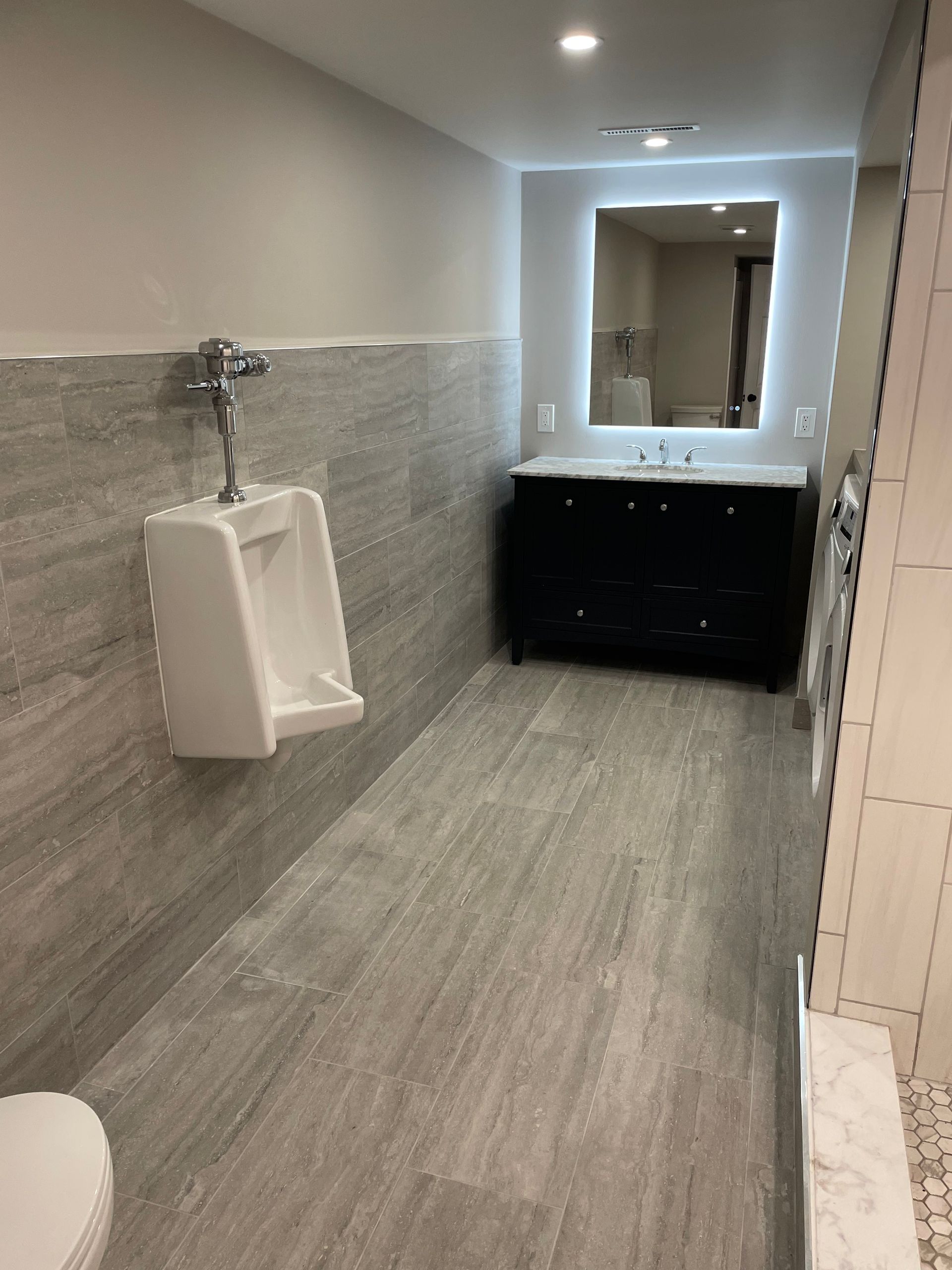 Modern restroom with gray tile, urinal, and black vanity sink under a lit mirror.