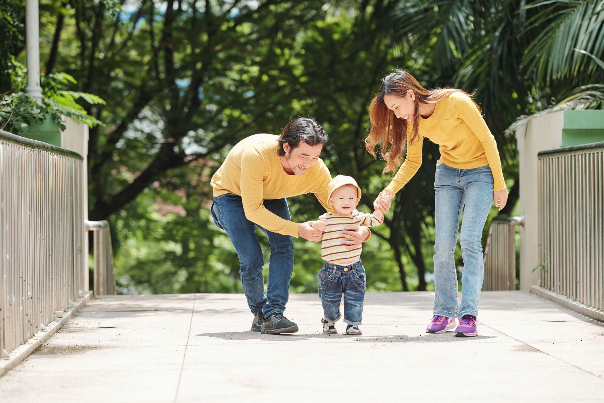 Parents in yellow sweaters help a toddler take steps on a bridge in a park.