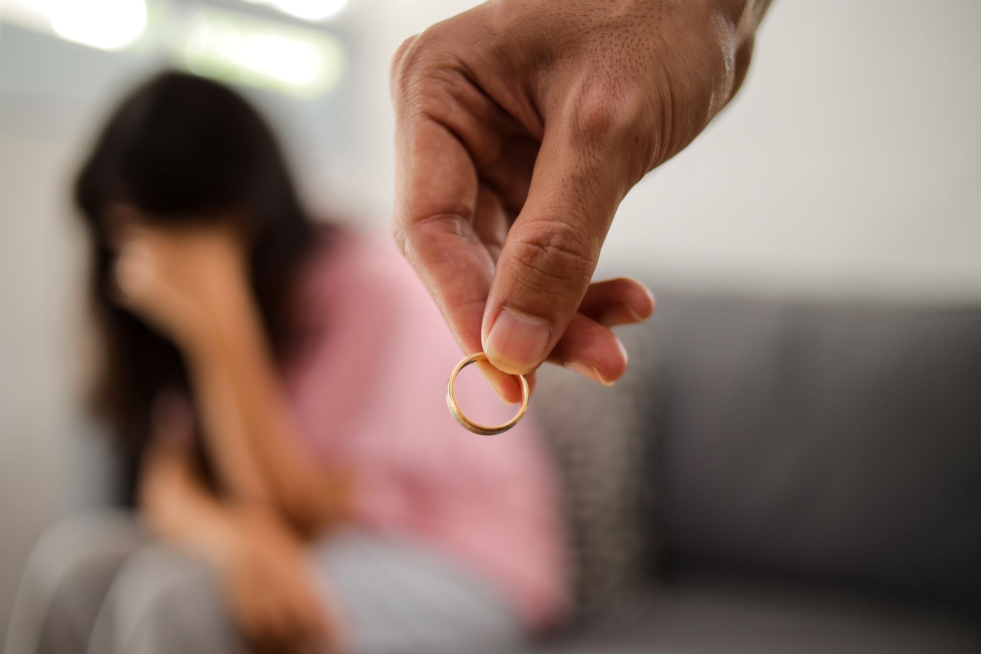 A hand holds a gold wedding ring in the foreground while a person sits blurred in the background with their head in hands.