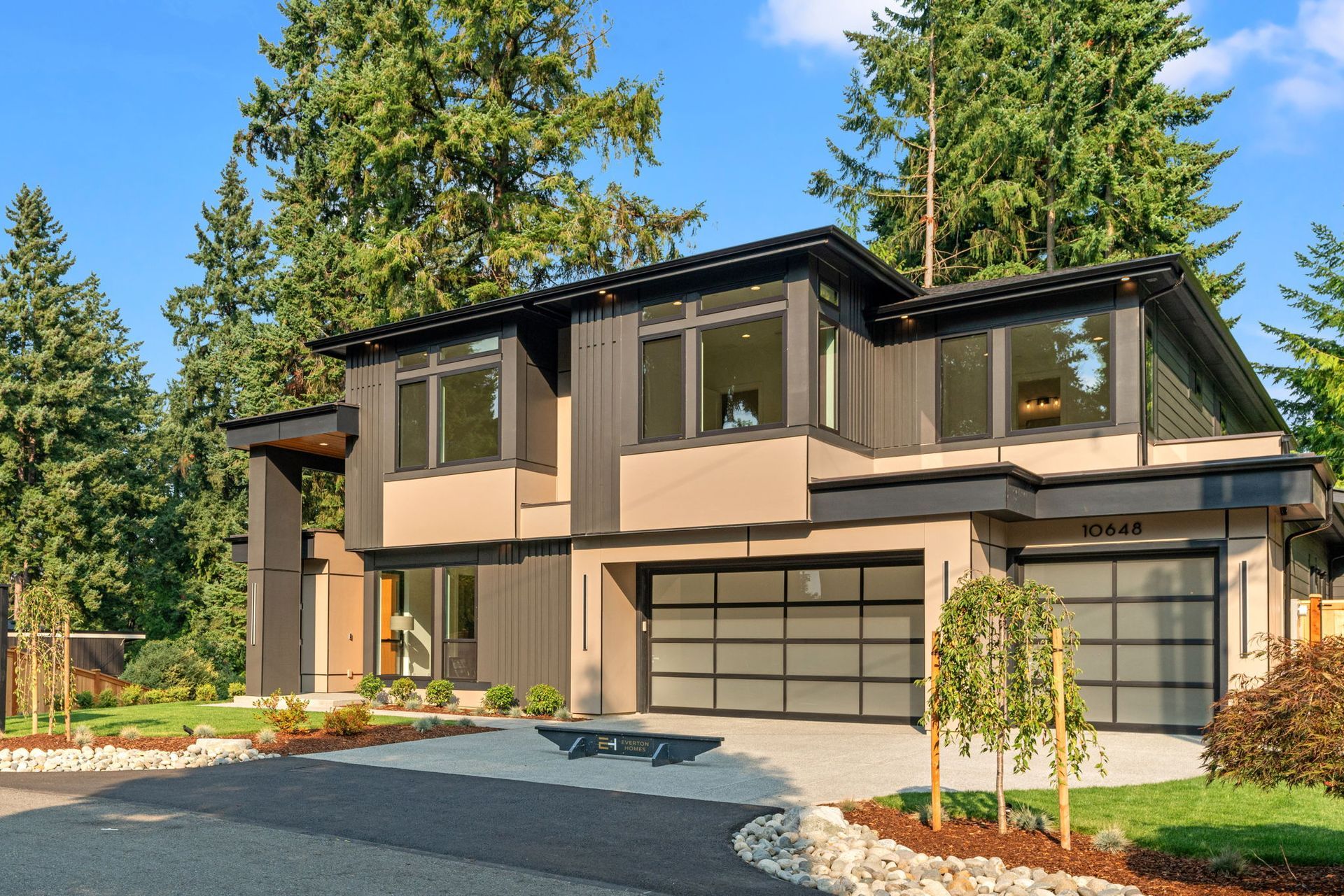 Modern two-story house with dark siding, glass garage doors, and large windows, set in a wooded area.