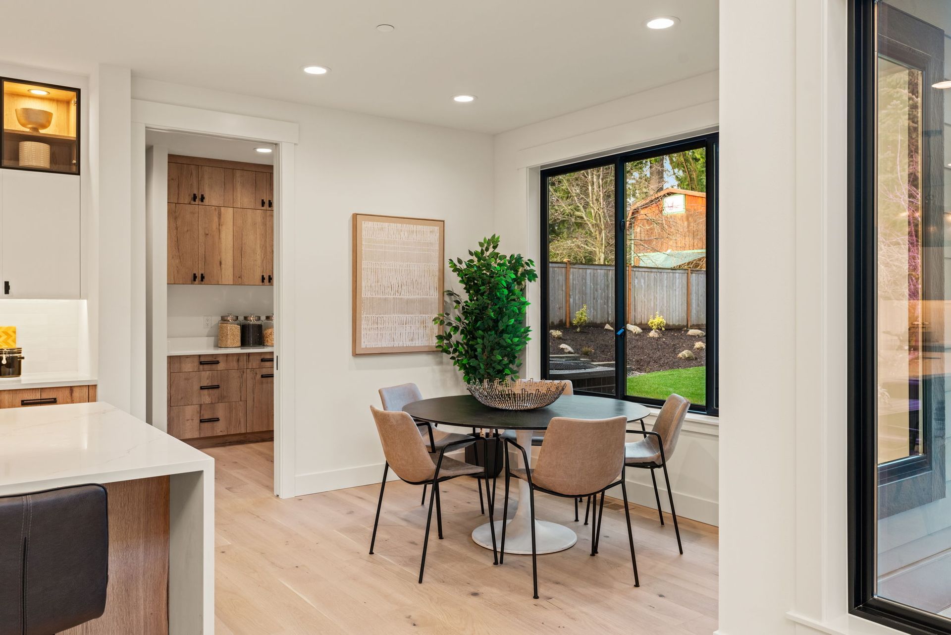 Dining area with round table, four chairs, and large window overlooking a yard; pantry and kitchen visible.