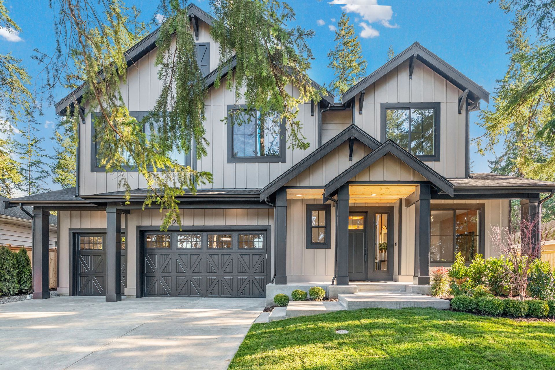 Two-story house with tan siding, black trim, and a gray garage door. Lush green lawn and trees in the foreground.