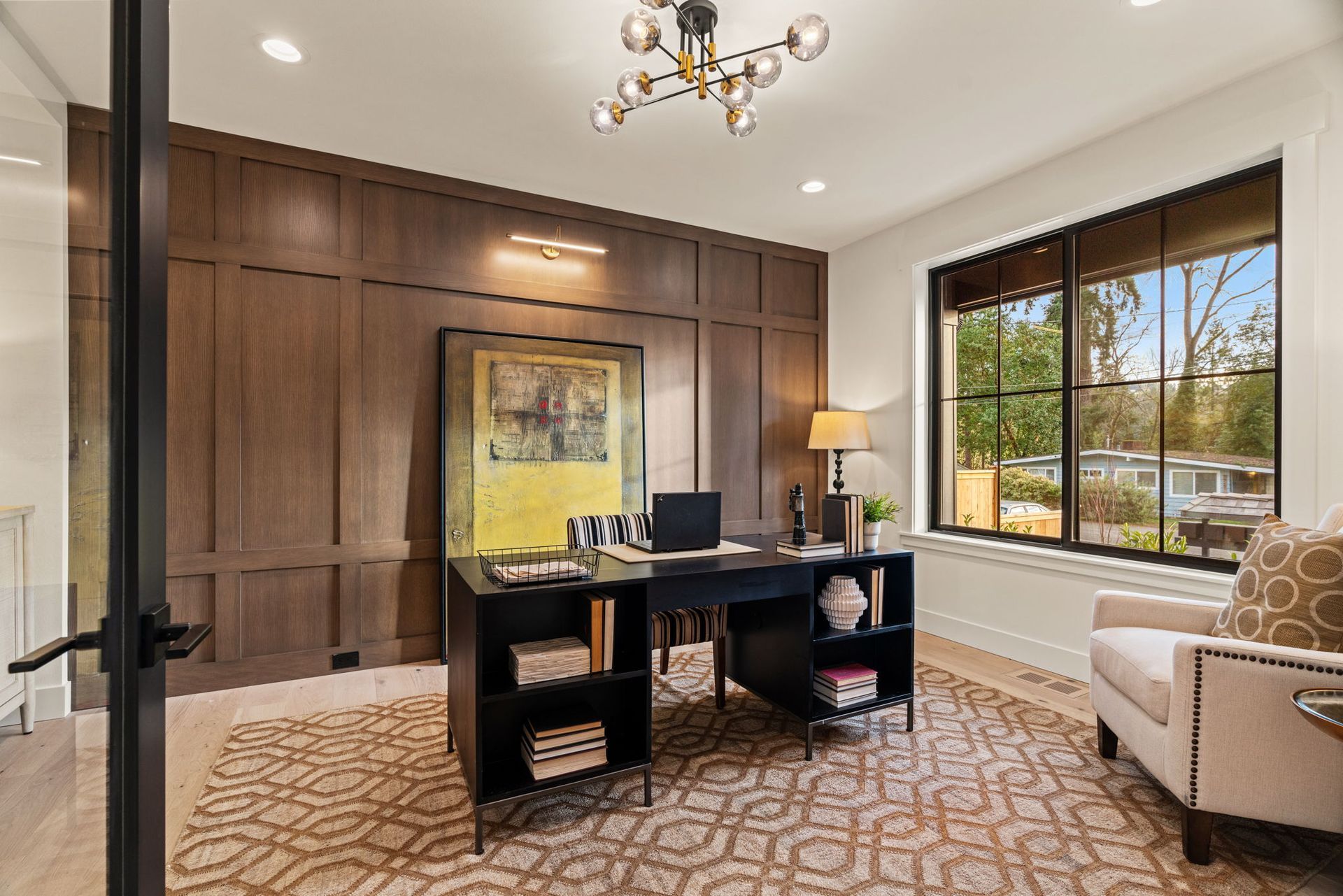 Home office with a dark wood paneled wall, black desk, patterned rug, and a window overlooking greenery.