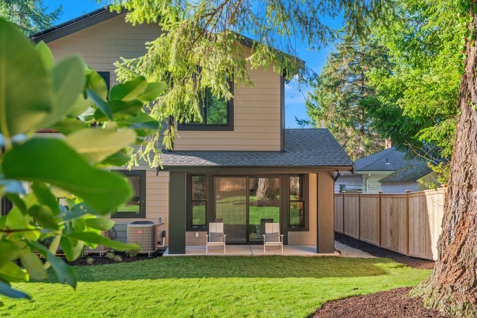 Two-story house with tan siding, large windows, and a patio, surrounded by a green lawn and trees.