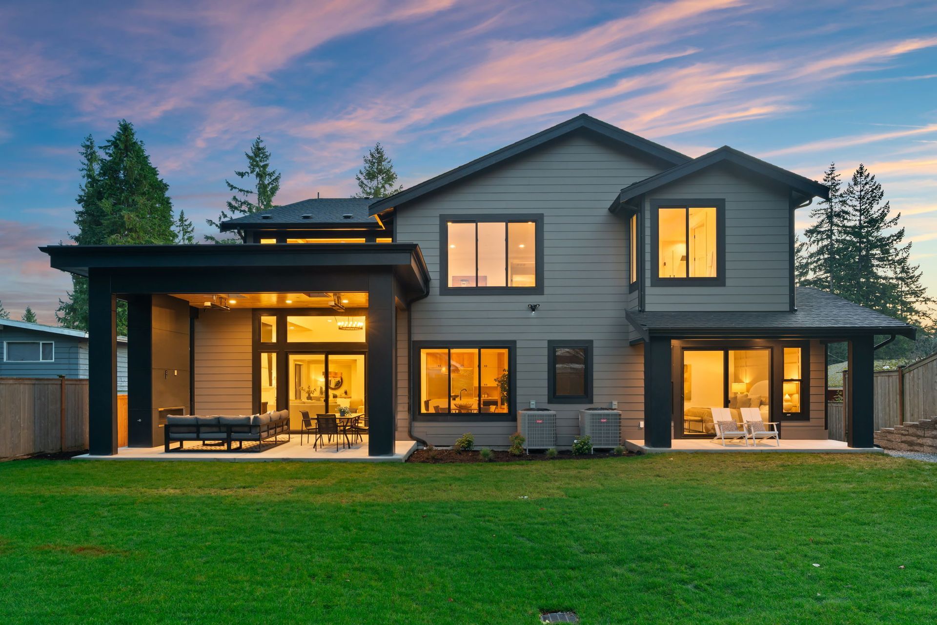 Rear view of a modern gray house with a green lawn and patio at sunset.