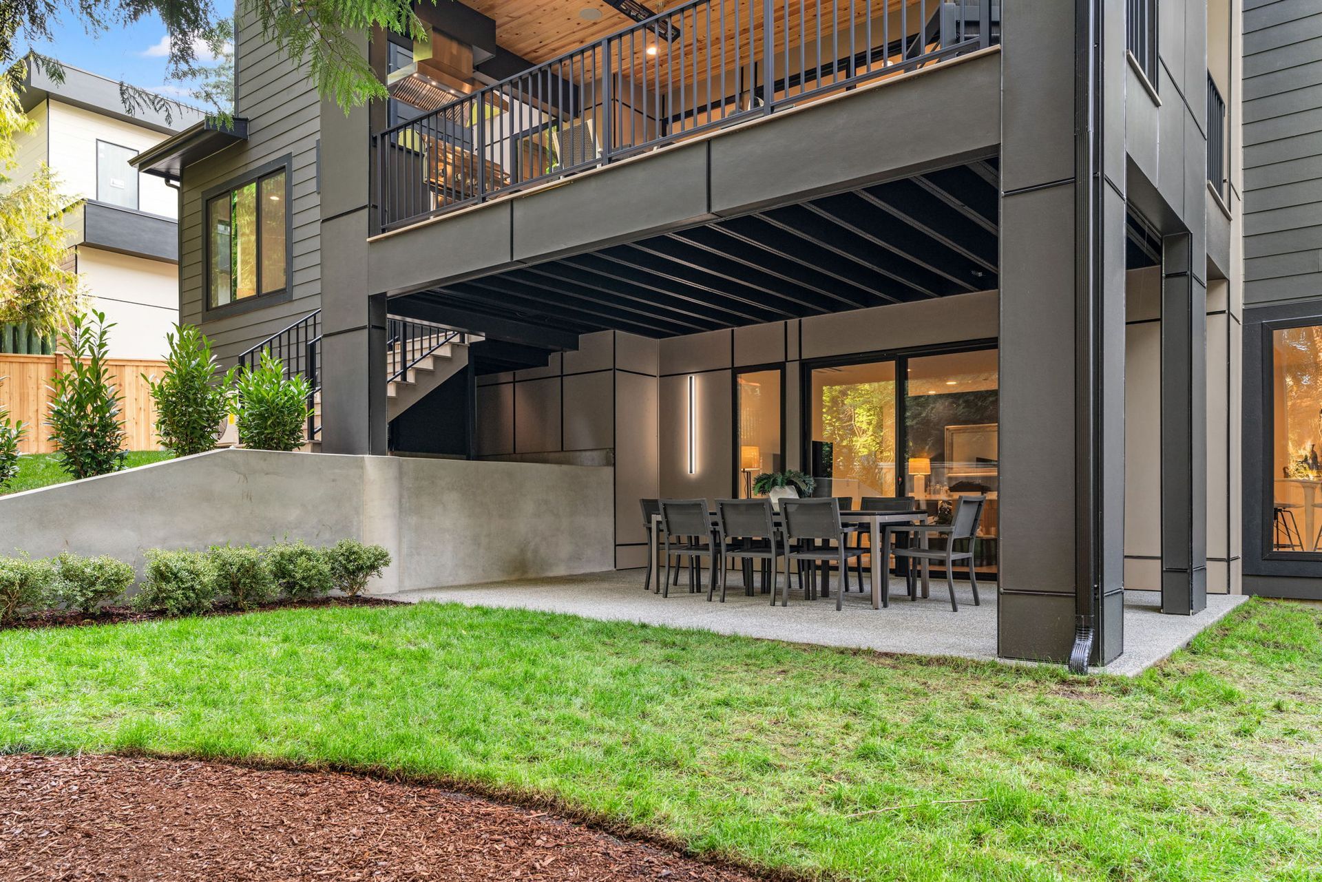 Modern home with a patio under a second-story deck, featuring a dining table on a grassy lawn.