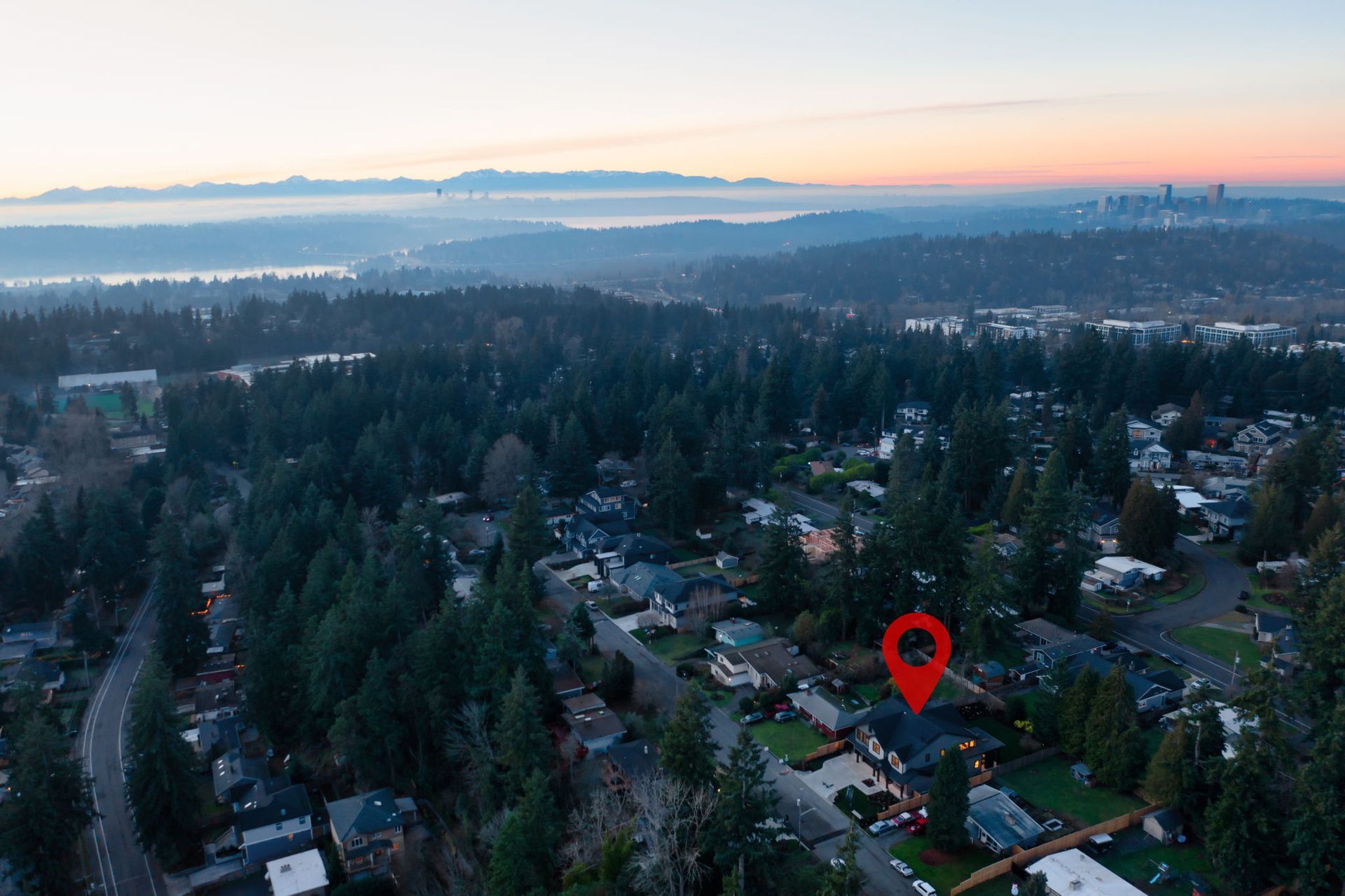 Aerial view of houses in a wooded area; red location marker pinpoints one house. Distant cityscape and mountains at dawn.