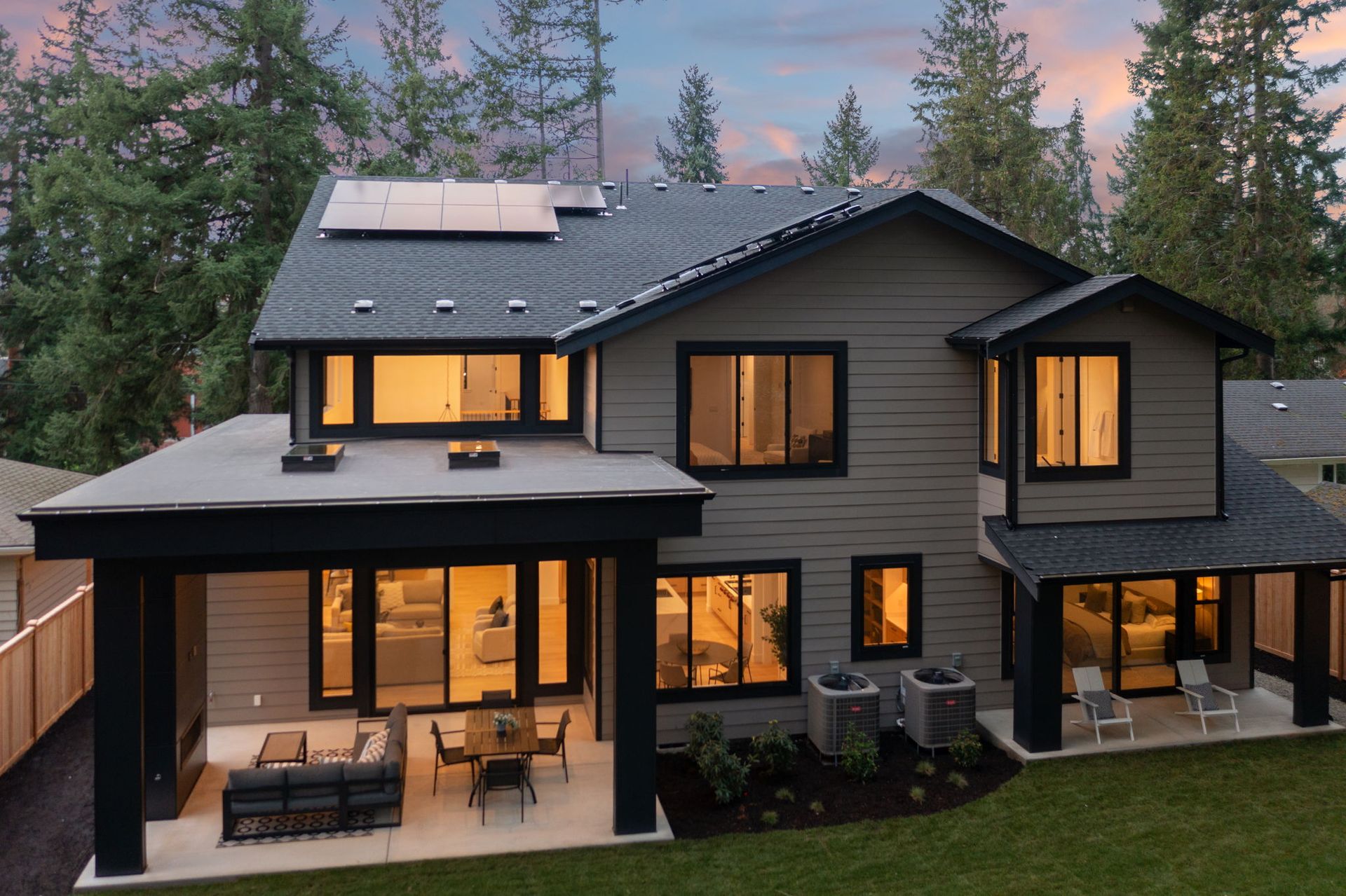 Modern two-story house with solar panels on roof and outdoor patio seating at dusk.