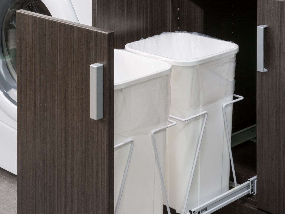 Two white trash cans are pulled out of a cabinet next to a washing machine.