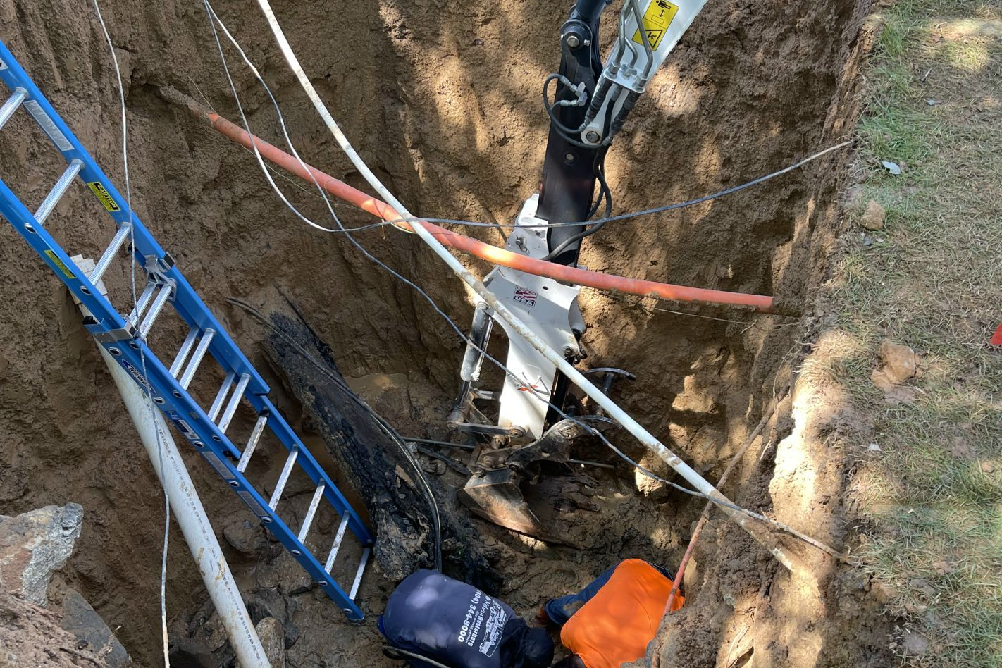 A worker in a trench repairs underground utilities. A ladder and cables are visible.