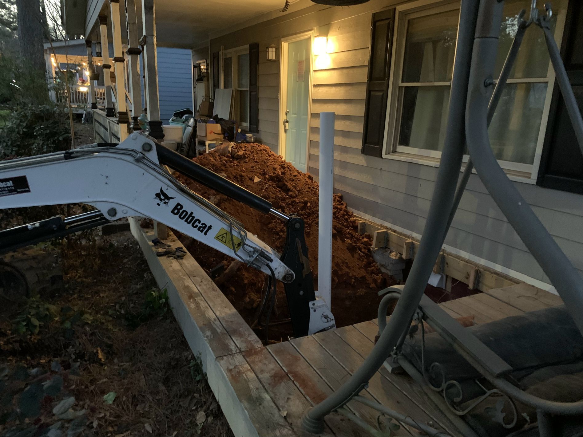 Bobcat excavator digging near a house, exposing a plumbing pipe. Brown dirt and gray siding visible.
