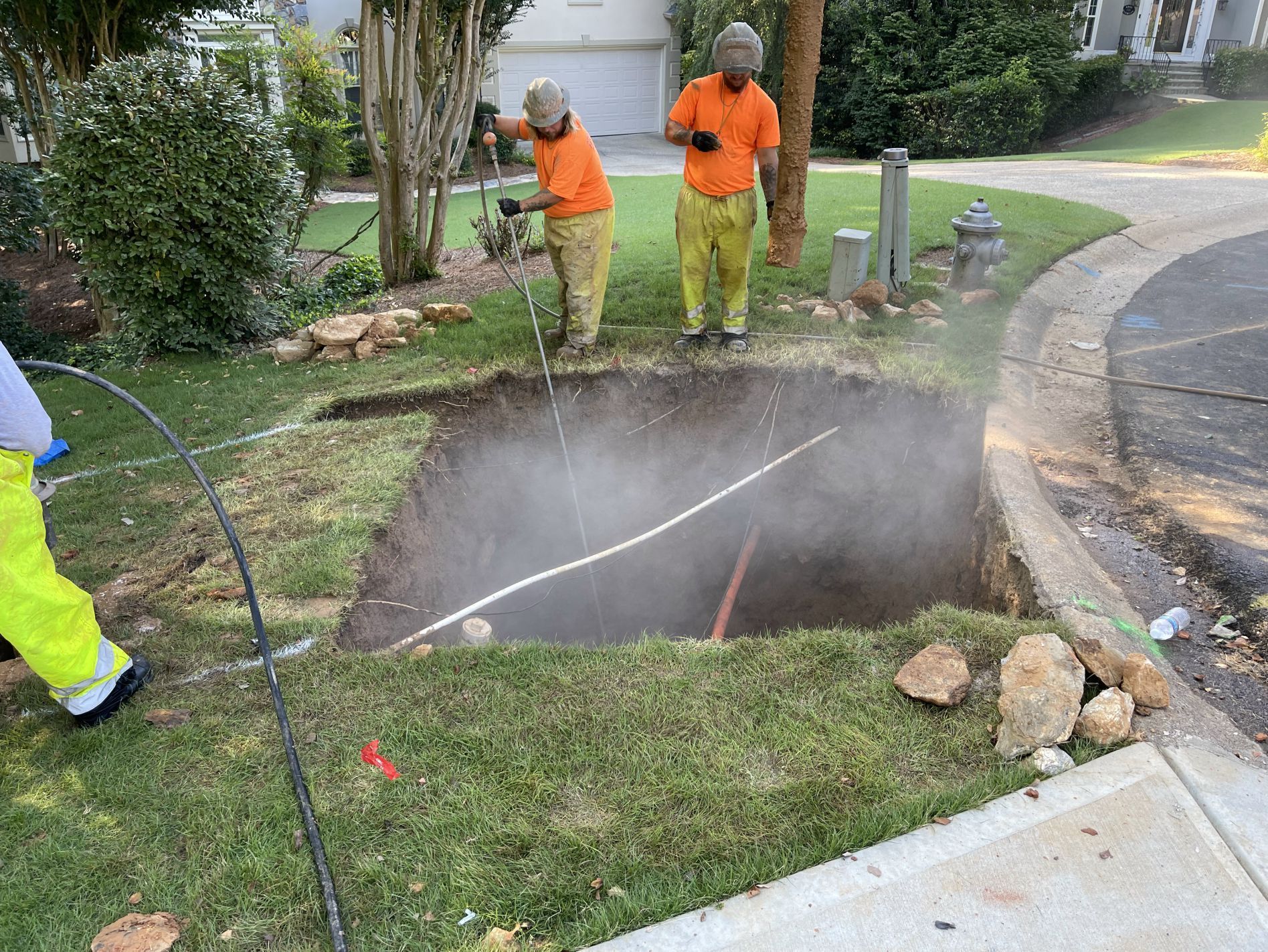 Utility workers in orange and yellow protective gear working in a hole in a grassy area, vapor rising.