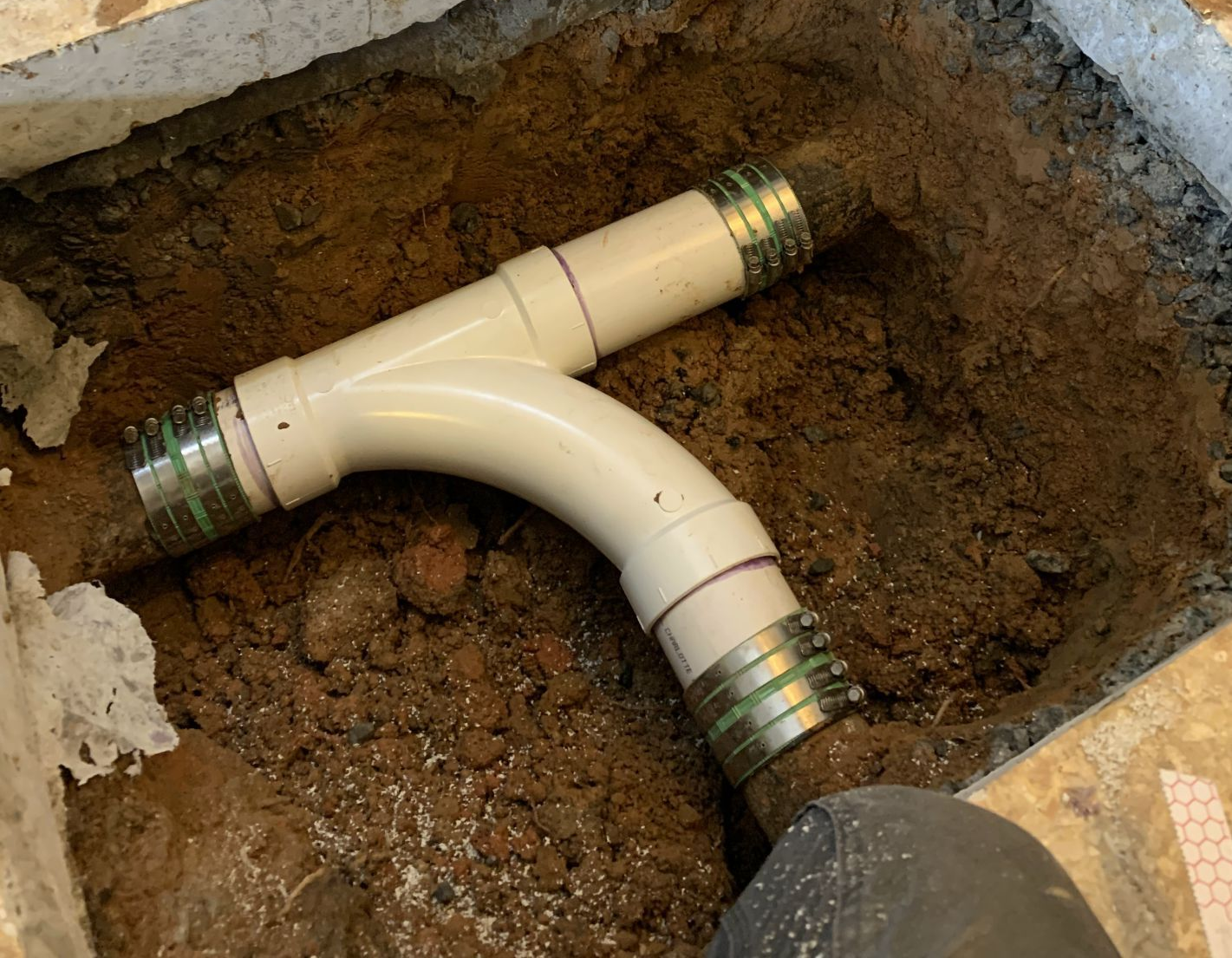 White PVC plumbing pipe with metal connectors, installed in a dirt-filled trench.