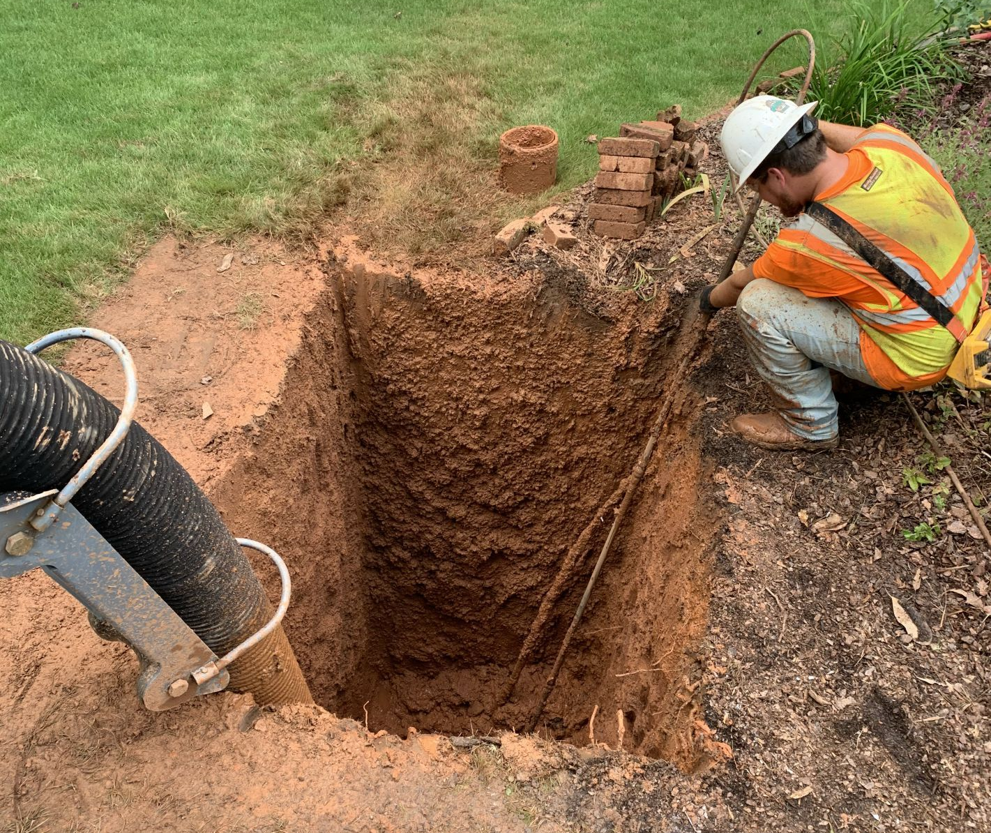 Worker in orange vest and hardhat uses a shovel in a dirt pit with a vacuum hose nearby.
