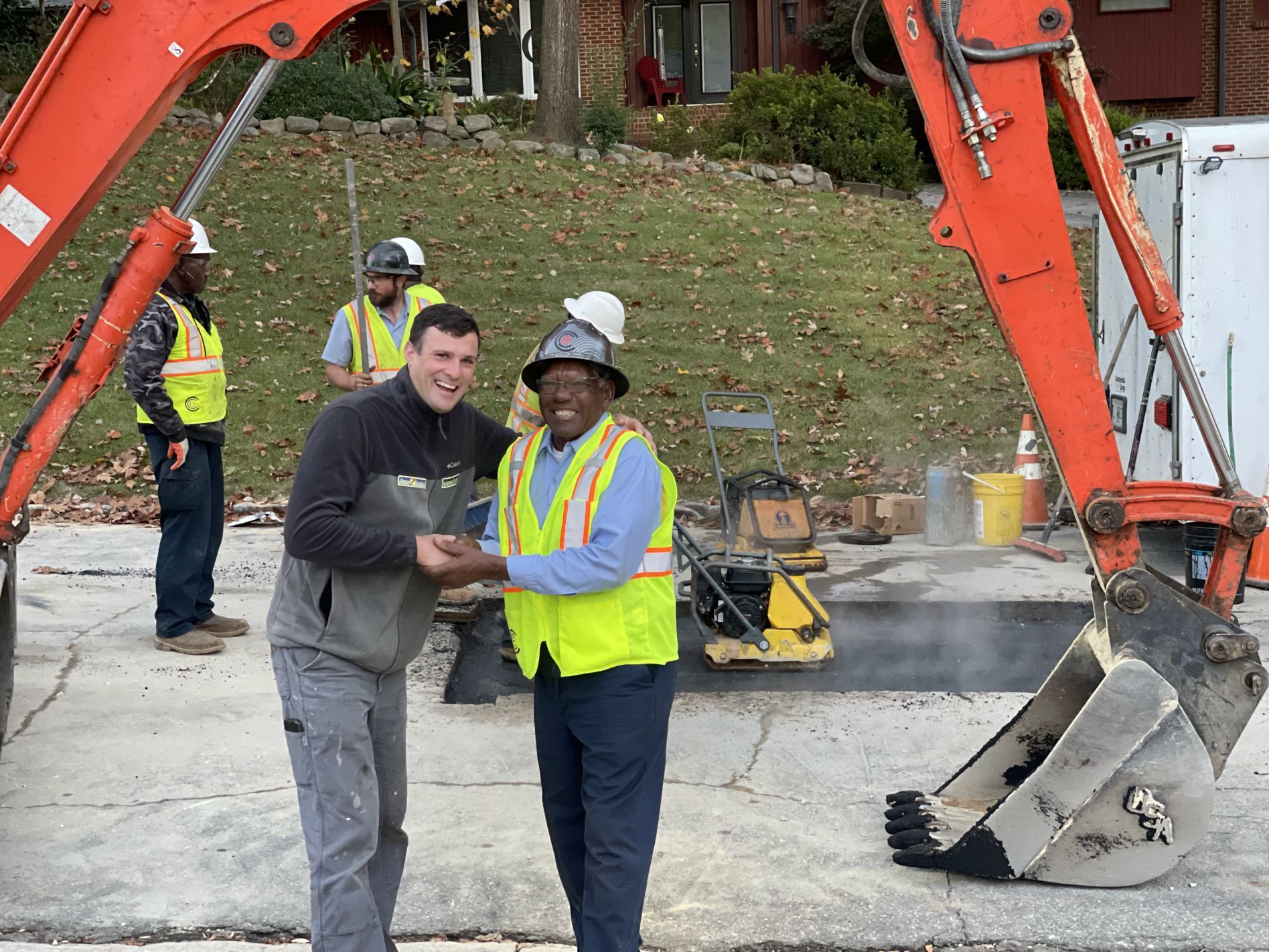Two men shake hands; construction workers in background; excavator on asphalt road.