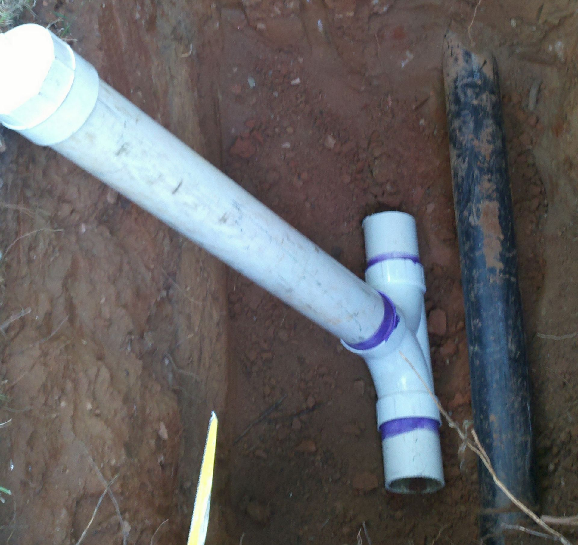 White PVC pipe connected to a black pipe in a dirt trench, with purple tape on joints.
