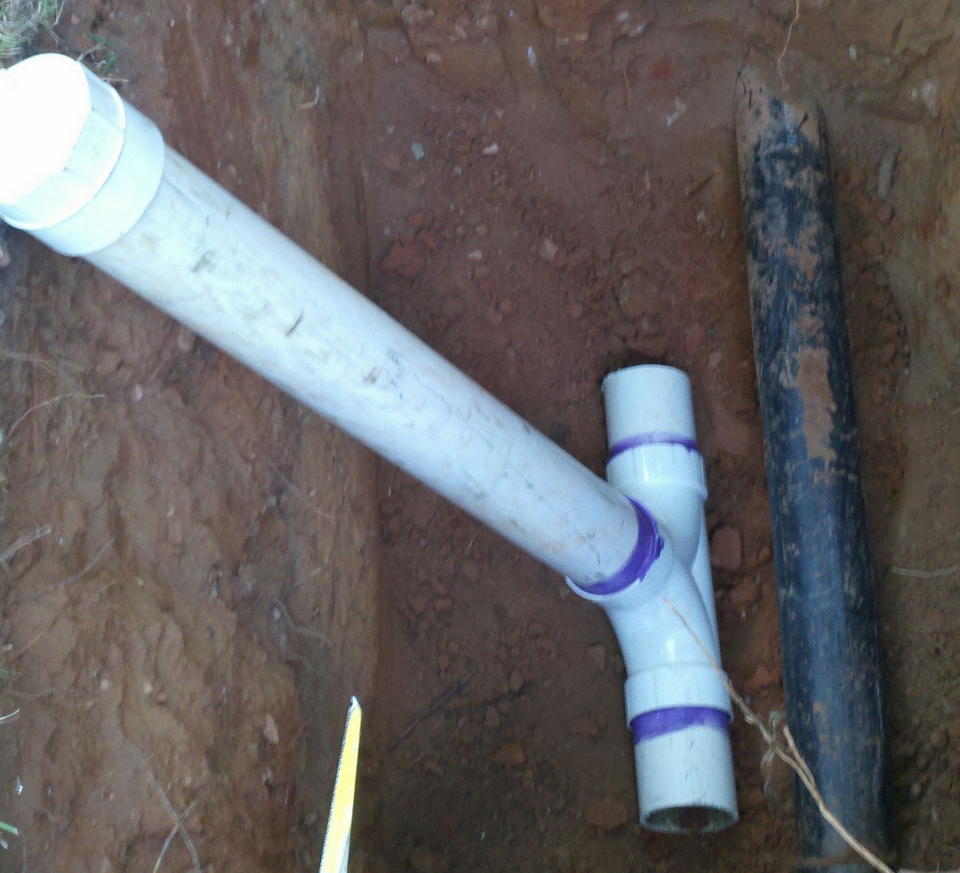 White PVC pipes and a black pipe connected in a dirt trench.