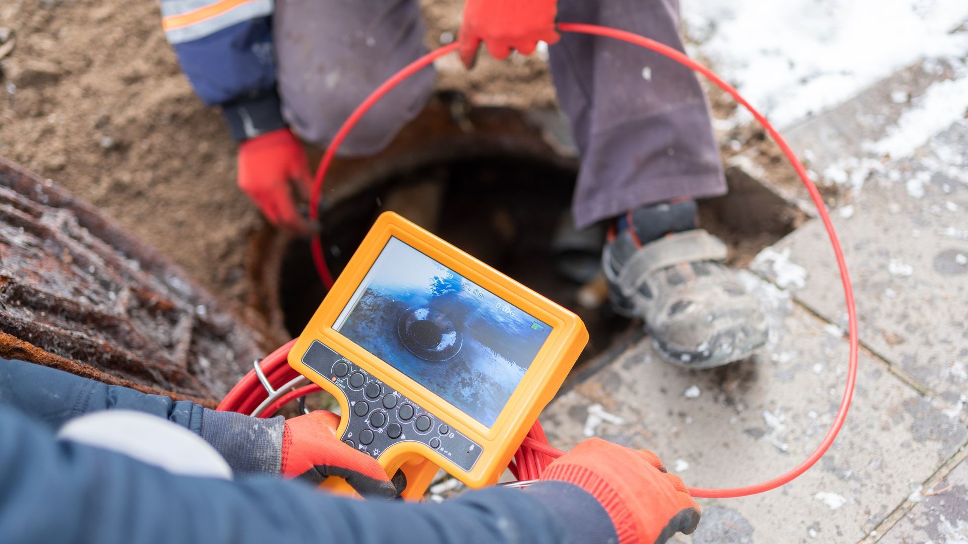 Workers inspecting a sewer with a camera monitor. Red gloves, orange screen, cable.