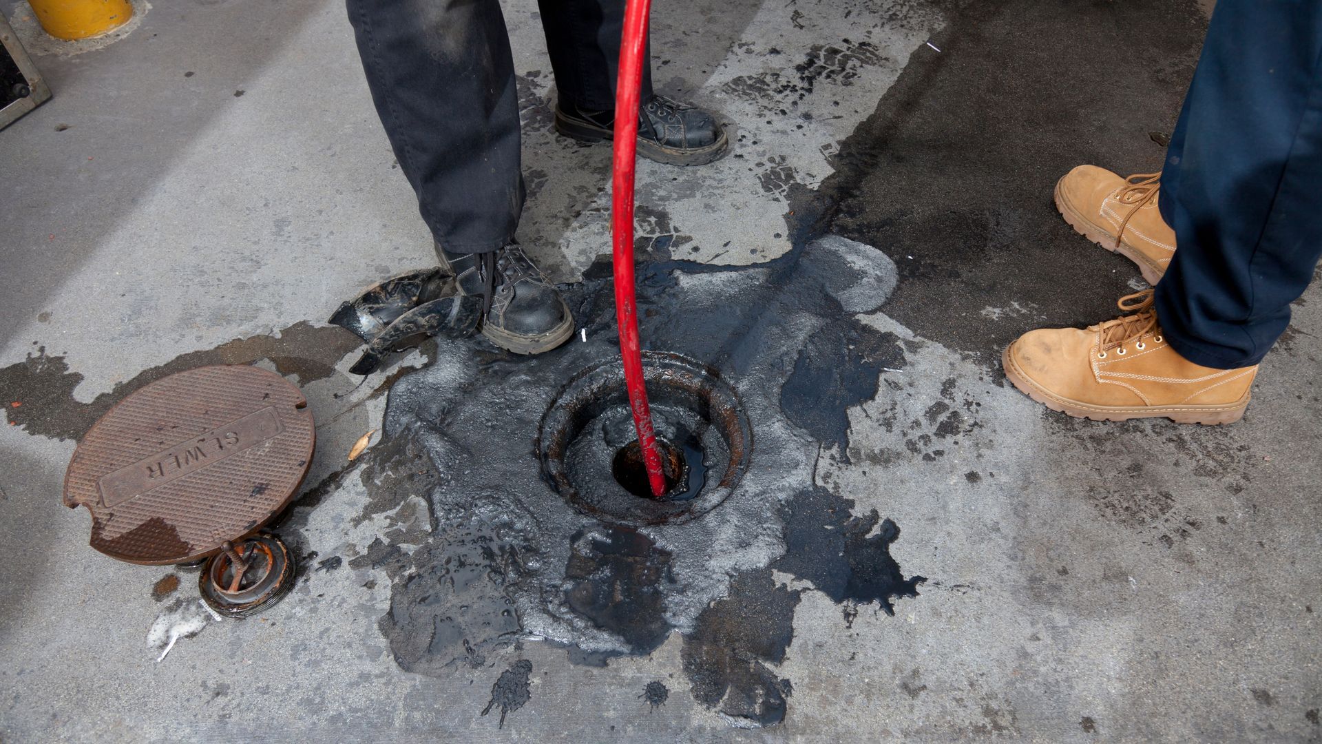 Workers using a red hose to clean a drain on a concrete surface, with a partially removed manhole cover nearby.
