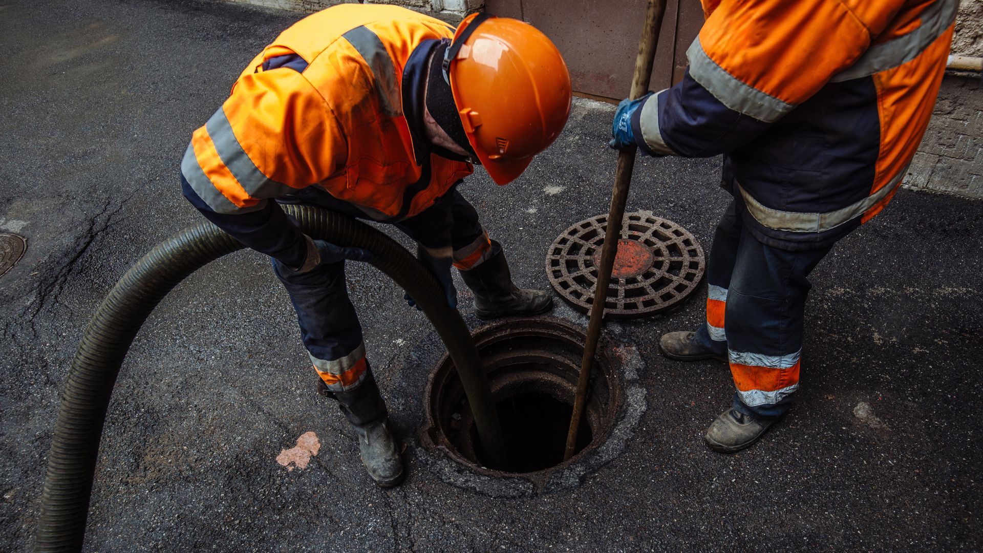 Two workers in safety gear cleaning a sewer drain in a street. One uses a hose, the other a rod.