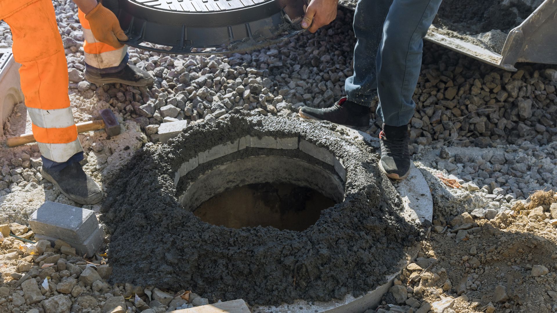 Workers applying cement around a manhole in a road with gravel; one wears an orange safety vest.