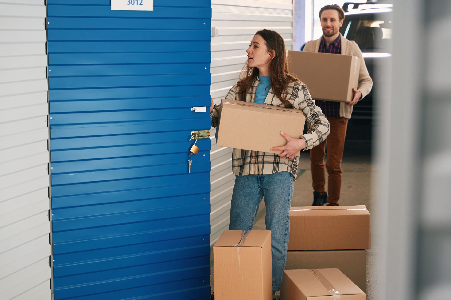 A young couple is holding cardboard boxes and opening the door of a blue-door self-storage rental.
