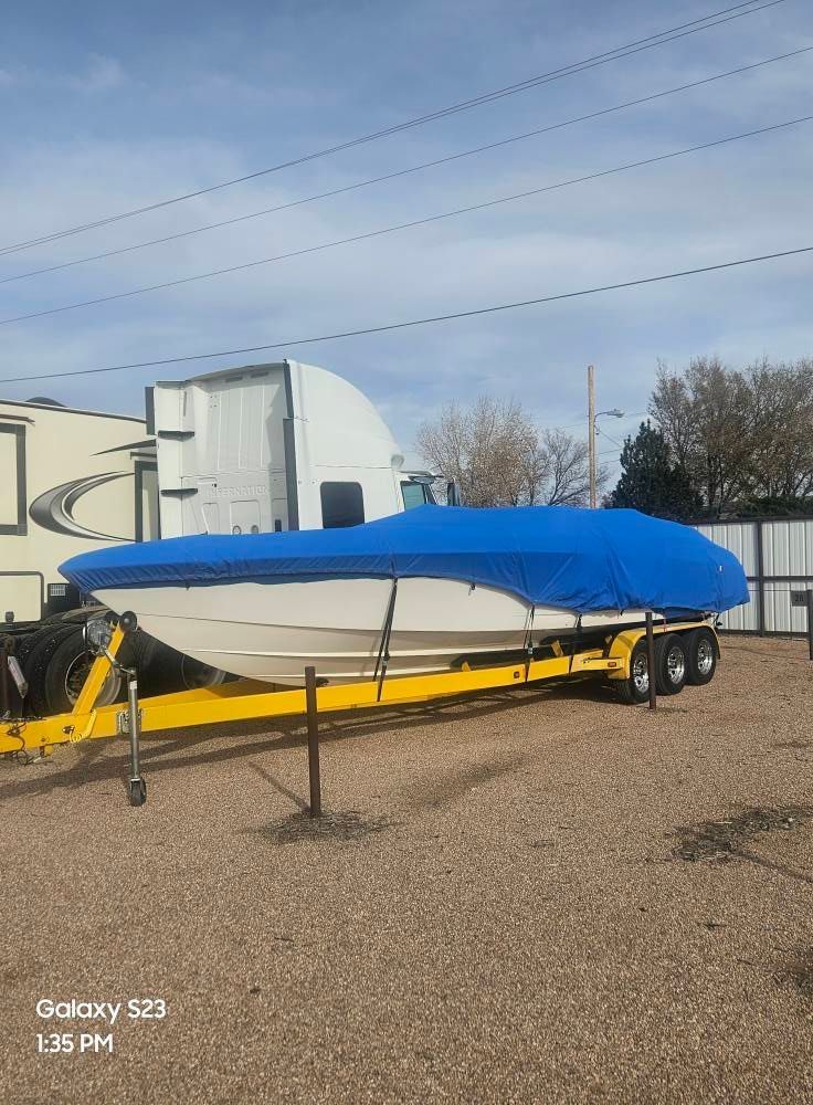 Boat on a trailer, covered with a blue tarp, parked outdoors on gravel. Yellow trailer in a sunny setting.