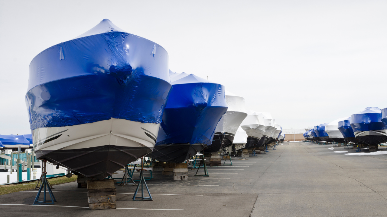 Boats covered in blue and white tarps on a paved lot, awaiting the boating season.